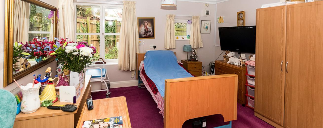 Tranquil Garden view bedroom, with assisted living featured single bed central to the room. Mass amounts of wooden furniture such as, wardrobe, two sets of chests of draws, bed side table and a comfortably placed mirror and television.