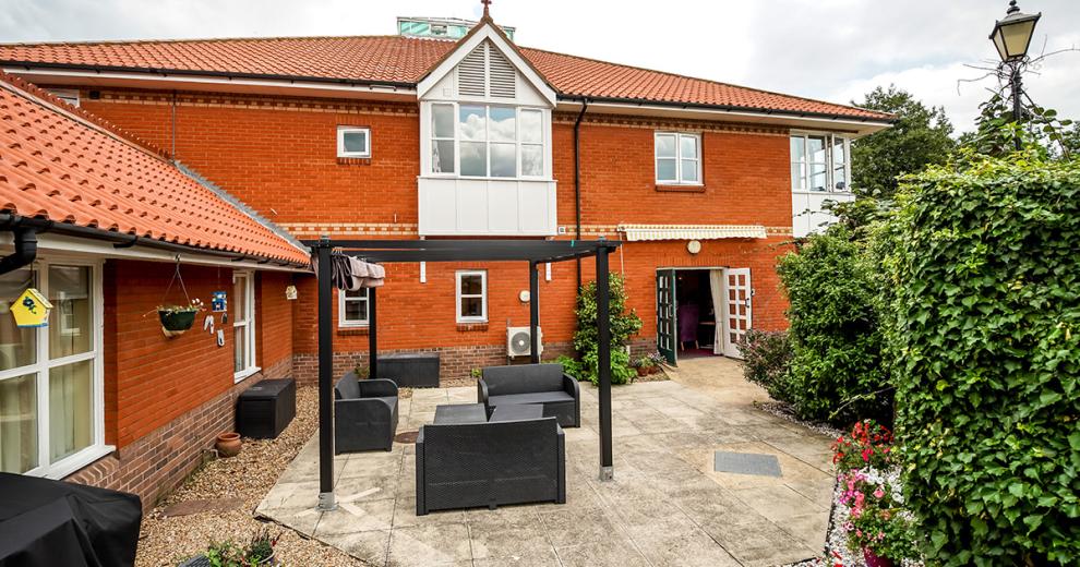 Commodious patio seating area with retracting cover for the pergola, covering three two-seater sofas and multiple tables. The garden is given privacy by large hedges and pretty flowerpots with no step access back through double doors into the property.