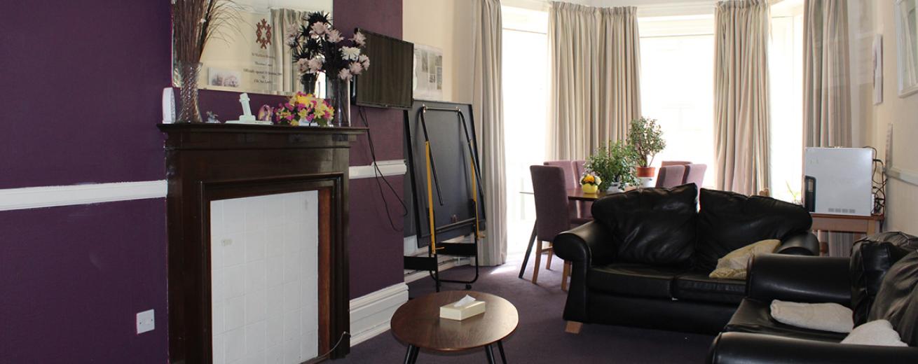 Well, lived in aubergine and cream communal living area. Made up of two black leather sofas, floral arrangements on the fireplace with large mirror hanging over.  And purple dining area to the back of the room in-front of the floor to ceiling bay windows.