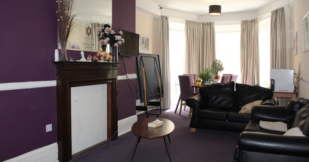 Well, lived in aubergine and cream communal living area. Made up of two black leather sofas, floral arrangements on the fireplace with large mirror hanging over.  And purple dining area to the back of the room in-front of the floor to ceiling bay windows.