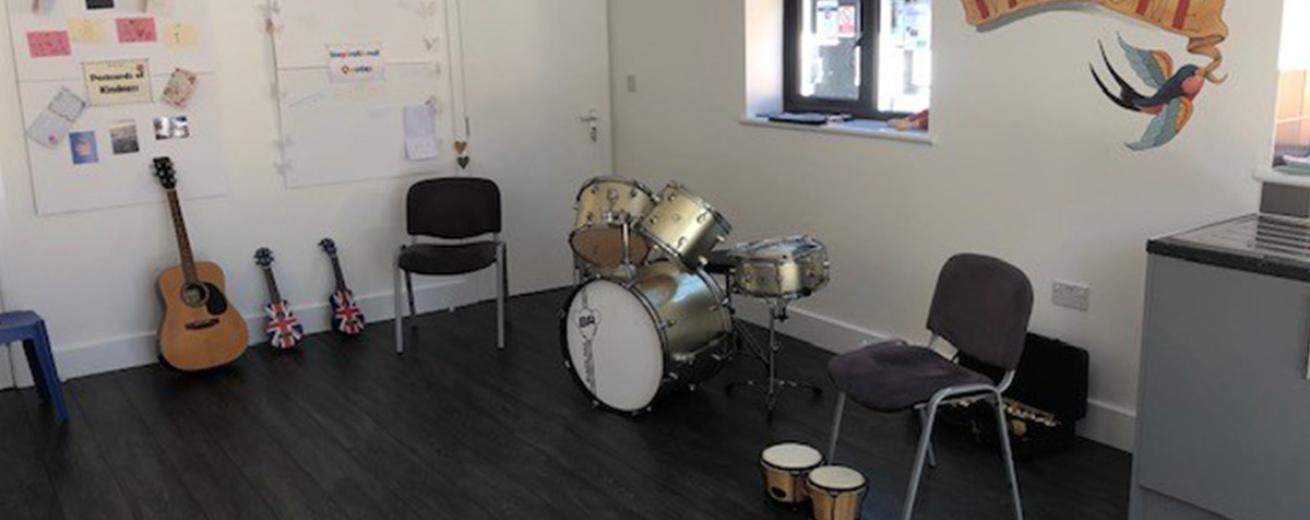 A naturally lit room with musical instruments including drum kit, a small wooden drum, a guitar and two small guitars. There are posters on the wall and a large welcome sign.