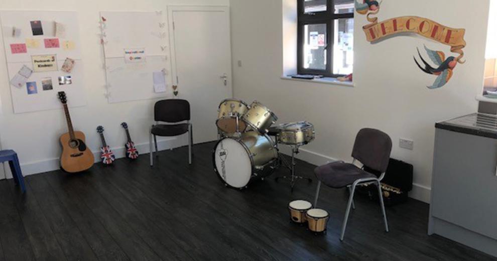 A naturally lit room with musical instruments including drum kit, a small wooden drum, a guitar and two small guitars. There are posters on the wall and a large welcome sign.