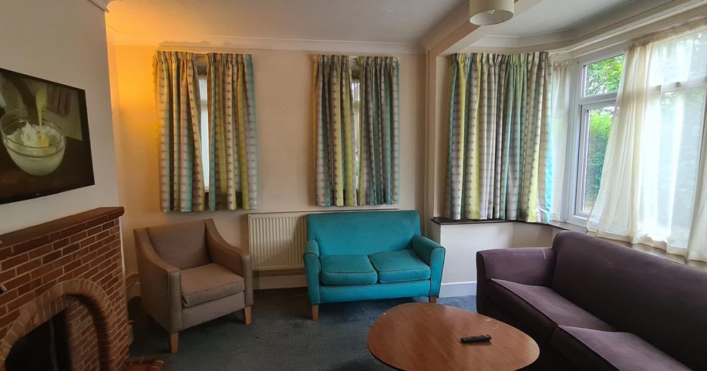 Minimalistic sitting area consisting of a deep purple three-person settee, a striking teal two-seater sofa and a beige curved armed single chair. Facing a brick built fireplace and wall mounted flat screen television.