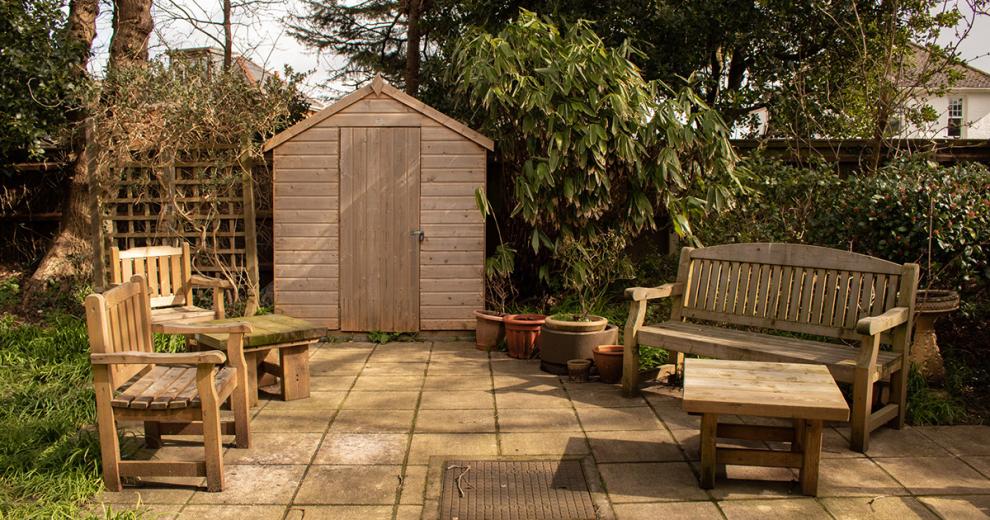 Composed, low-maintenance patio seating area with bench and chair availability as well as small matching wooden coffee style tables. All in front of a secure shed nestled into the large vegetation.