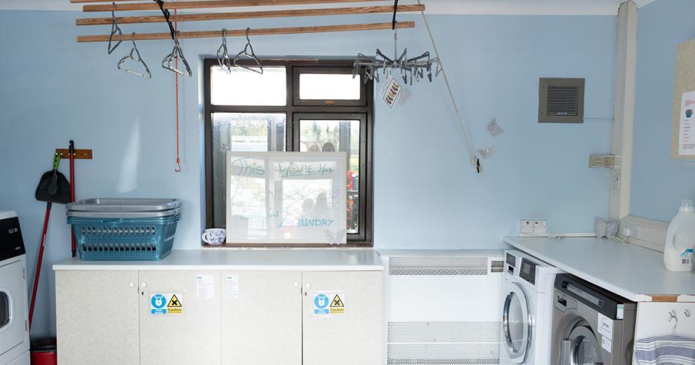 Pale blue laundry room, housing multiple washers, dryers and wash baskets. As well as an overhead drying rack with hangers and pegs.