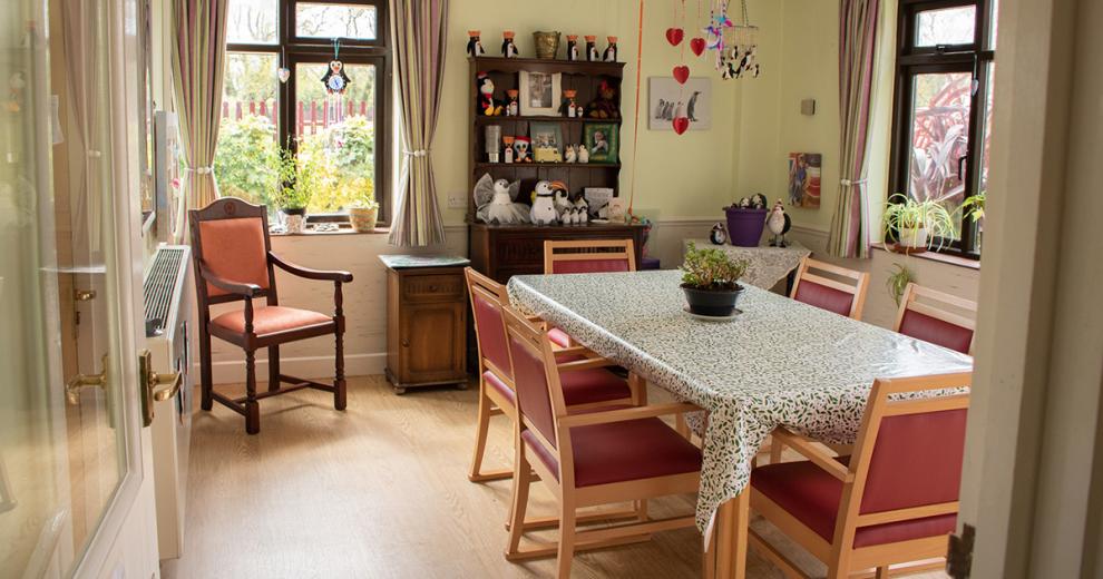Homely, dining room area with bulky wooden furniture, red upholstered dining chairs and patterned tablecloth.