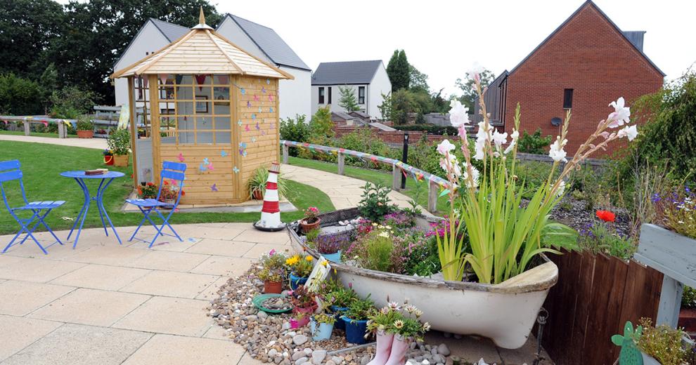 Lively flourishing garden with a large boat planter featuring an array of colourful mature plants. Surrounded by novelty wellington boot plant pot and pastel-coloured potted wildflowers. Petite stand-alone wooden garden room, with personal touches and a bold royal blue metal garden seating set.