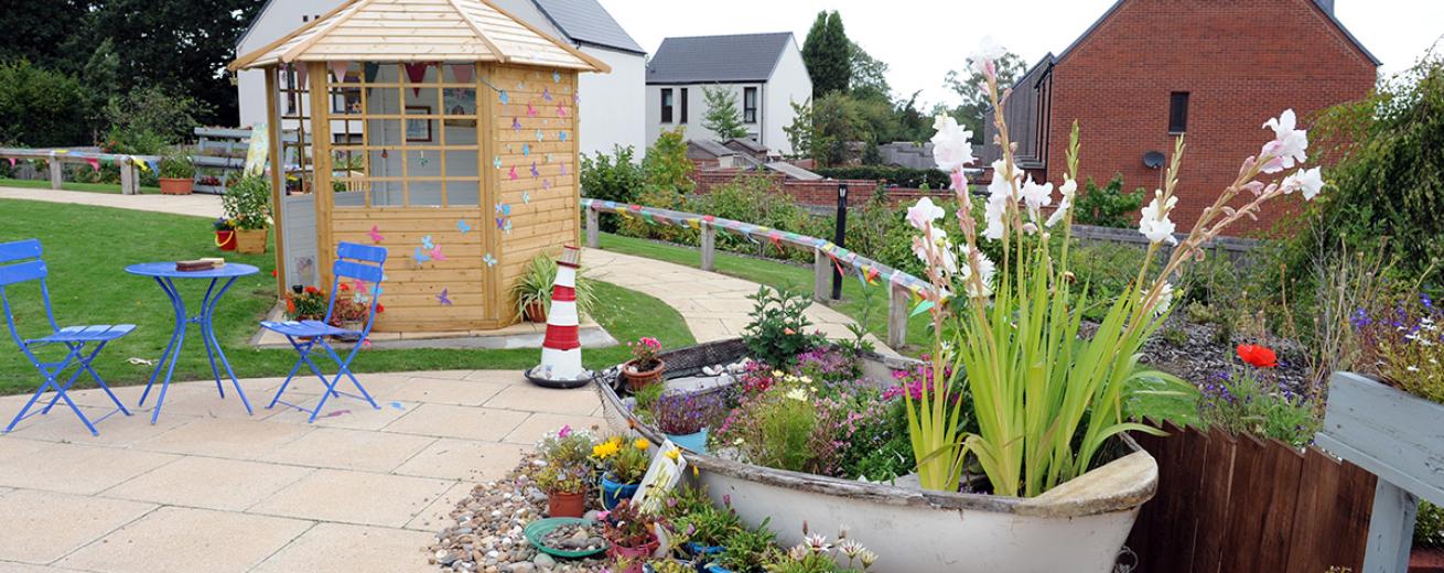 Lively flourishing garden with a large boat planter featuring an array of colourful mature plants. Surrounded by novelty wellington boot plant pot and pastel-coloured potted wildflowers. Petite stand-alone wooden garden room, with personal touches and a bold royal blue metal garden seating set.