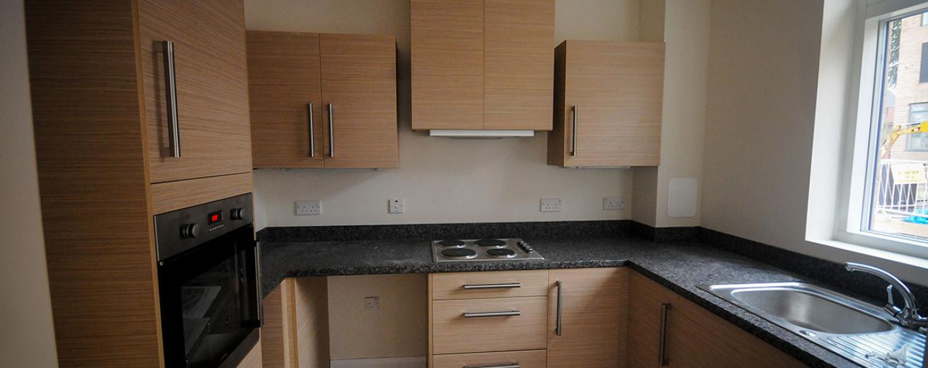Humble sized kitchen area, consisting of midtone beige kitchen units with sturdy silver handles. Integrated wall mounted oven and counter fitted electric hob.