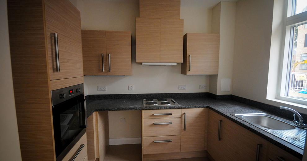 Humble sized kitchen area, consisting of midtone beige kitchen units with sturdy silver handles. Integrated wall mounted oven and counter fitted electric hob.