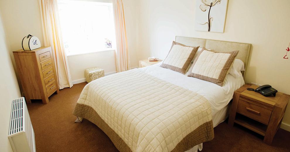 A relaxing bedroom with a centrally placed double bed with cream headboard, white bedding a cream and brown throw over and matching decorative cushions. Two oak wooden side tables sit either side of the bed and a complementing oak chest of draws are angled in the corner, leaving plenty of floor space to manoeuvre around.