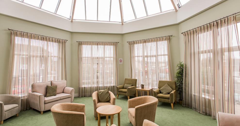 The sizeable extension floods natural light into the seating area. The angled glass panelled roof and oversized windows on every exterior wall. The furniture consists of four light brown coloured bucket armchairs surround a small coffee table in the middle of the room, two olive green plush arm chairs tuck neatly into the window and a two person lilac sofa sits opposite them.