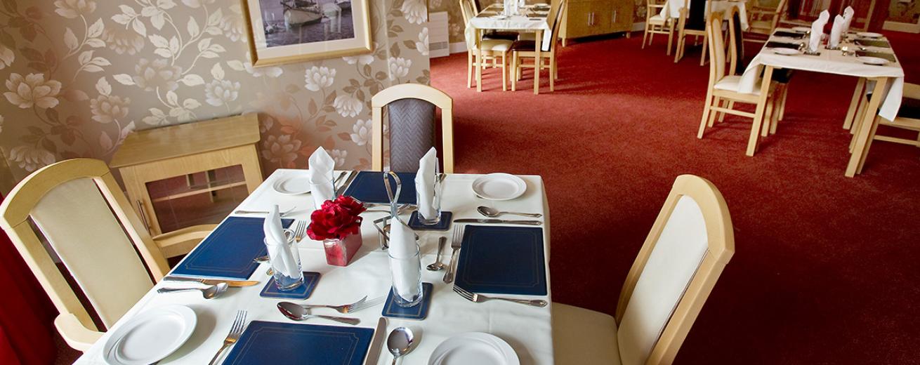 The spacious dining room is home to multiple dining tables dressed in white linen cloths and matching chairs with cream and aubergine coloured leather upholstery. The tables are dressed with leather placemats, silver cutlery.