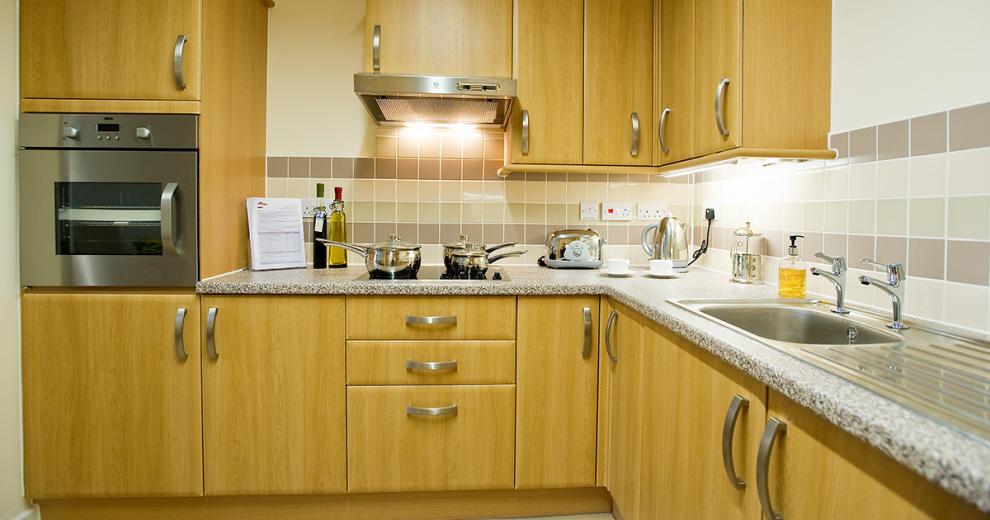 A warm and inviting kitchen area, featuring oak wood cabinetry with white painted walls and earthy brown and grey toned wall tiles. The wall mounted oven, extractor fan and sink are all in chrome which reflects the light perfectly. The room is finished with high end silver cooking utensils along the worktops.