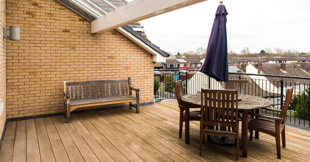 A reasonably sized wooden decked balcony seating area. Home to a weathered circular brown wooden outdoor dining set and a wooden bench up against the wall.