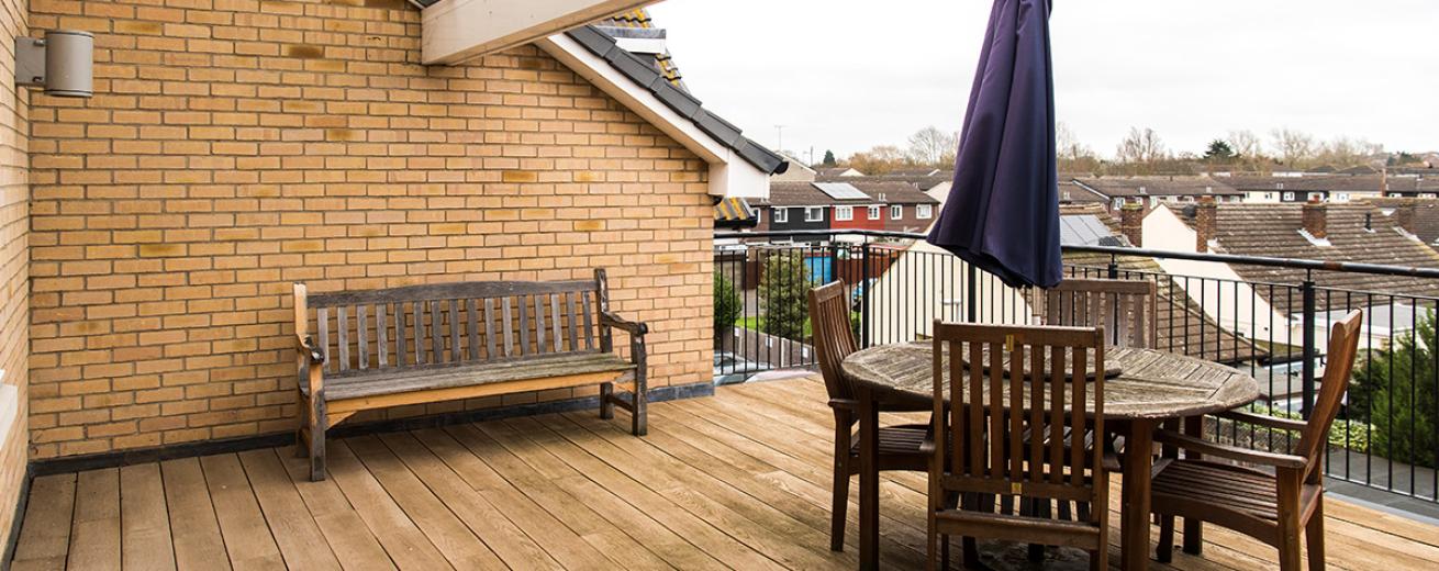 A reasonably sized wooden decked balcony seating area. Home to a weathered circular brown wooden outdoor dining set and a wooden bench up against the wall.