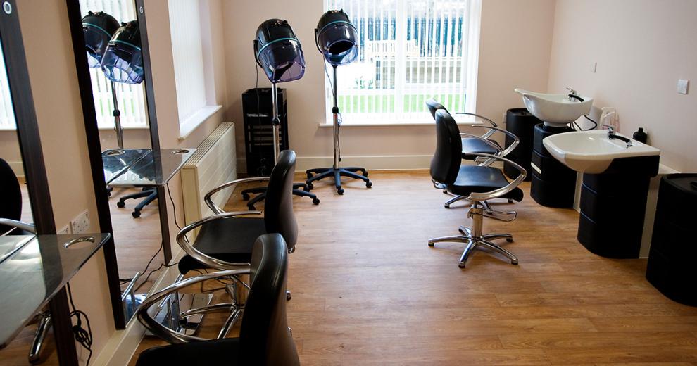 A classically designed hair salon area. Two large, elongated mirrors with thin wooden framing are the main feature of the room. Two plush black styling chairs are located in front of the mirror. Two hair washing basins sit opposite the styling area.