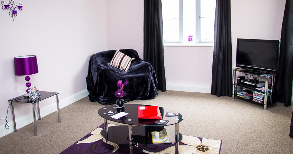 Subtle purple seating area, with large velvet covered single seater chair, coordinating black glass coffee table, television unit and side table. With deep purple and floral decorations.