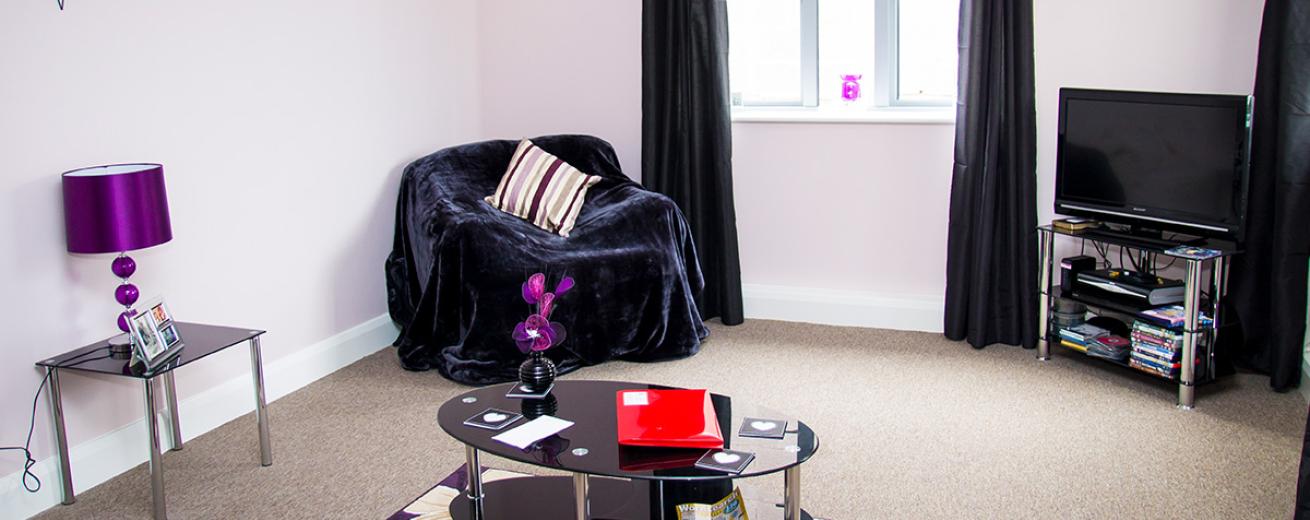 Subtle purple seating area, with large velvet covered single seater chair, coordinating black glass coffee table, television unit and side table. With deep purple and floral decorations.