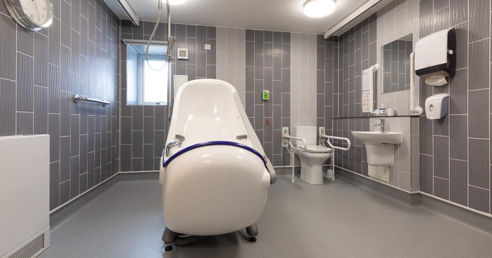 State of the art accessible wet room, with brand new bath and hoist system and plenty of space for comfort. A white ceramic toilet with fitted hand railings and ceramic wall mounted sink. In a multi-tone grey tiled decorative pattern and grey resin flooring.