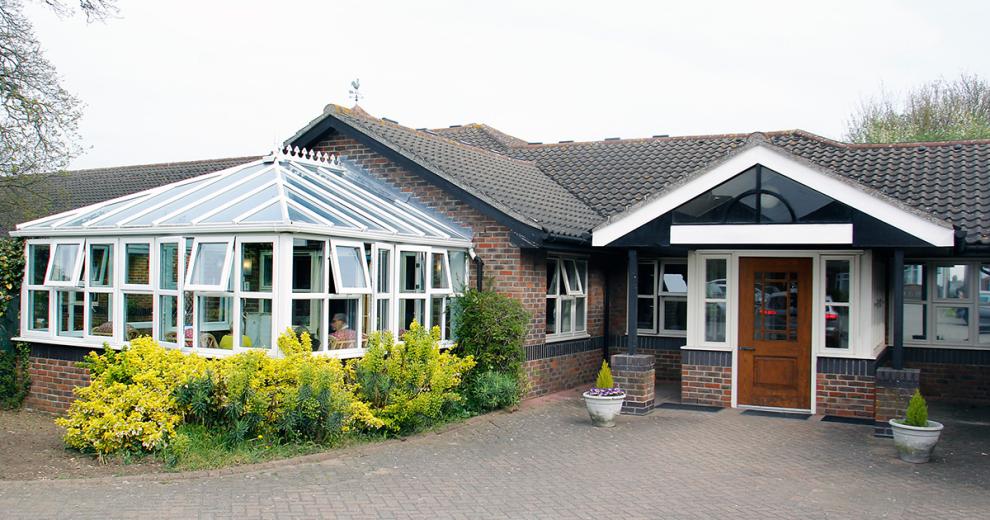 An inviting bungalow building, with multiple white PVC windows to allow masses of natural light. The building has an impressive conservatory extension with beautiful shrubbery growing around it.