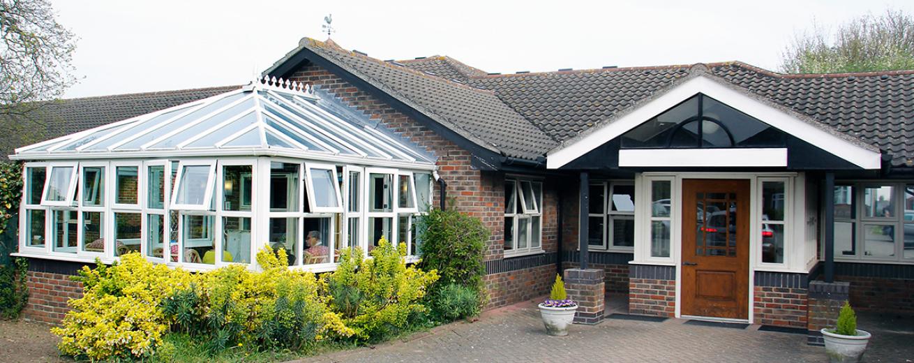 An inviting bungalow building, with multiple white PVC windows to allow masses of natural light. The building has an impressive conservatory extension with beautiful shrubbery growing around it.