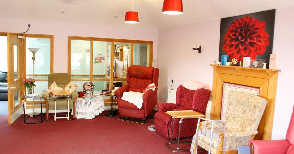 A broad seating area, with glass panelled wall looking out into the corridor, Featuring a mixed bunch of seating options, from a red leather electric armchair, red and white patterned reclining chair, and sturdy traditional patterned armchairs.