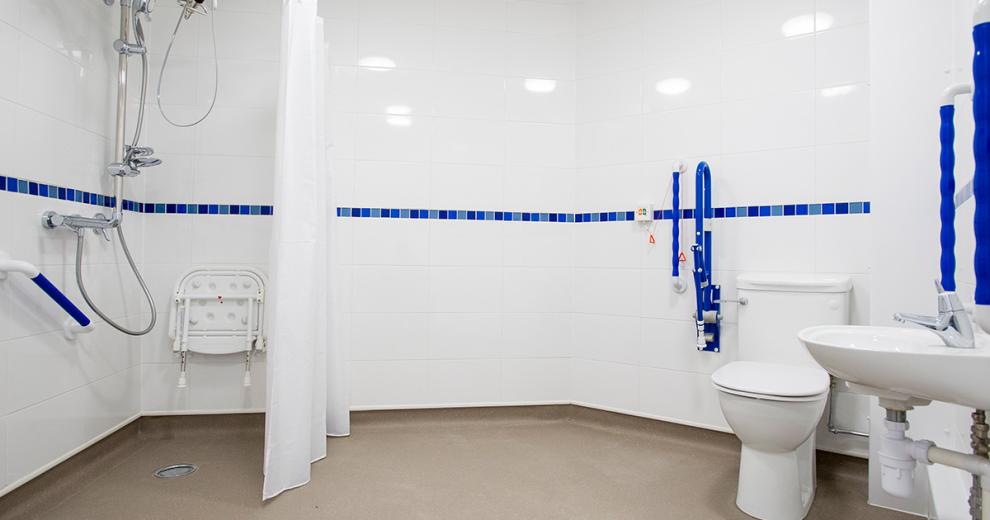 Sparkling clean and fresh, pure white wetroom with blue toned central strip and comfortable blue grips to help facilitate with accessibility. The shower area is sectioned off with a fabric curtain and features a fold up shower chair.