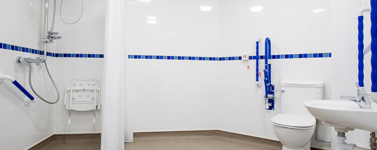Sparkling clean and fresh, pure white wetroom with blue toned central strip and comfortable blue grips to help facilitate with accessibility. The shower area is sectioned off with a fabric curtain and features a fold up shower chair.