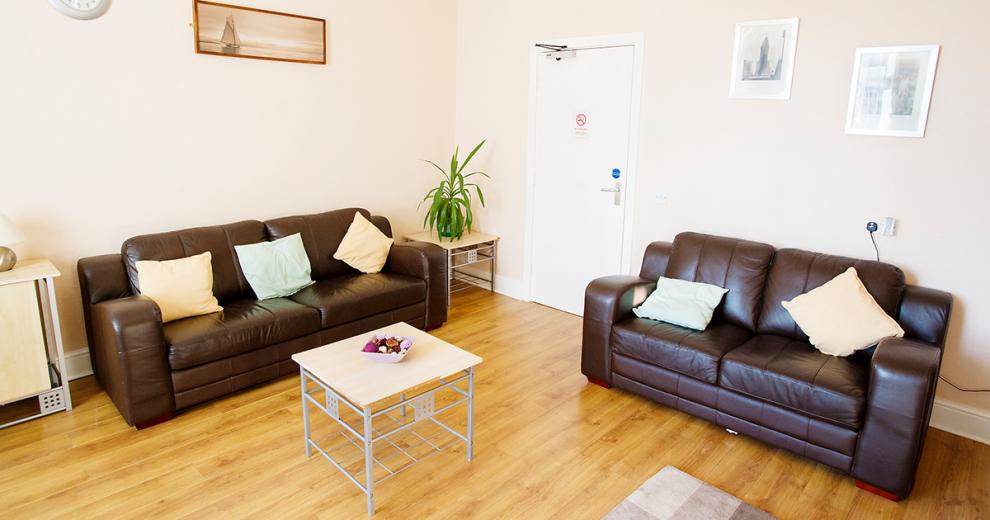 A fresh seating area, providing two large brown leather sofas and light toned cushions, with laminate flooring and a few small, squared coffee tables.