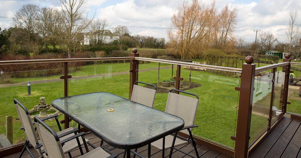 The balcony spans into a right angle. The dark wooden decking and glass panels give a seamless view into the garden area. A four person metal and glass outdoor dining set are tucked into the corner.