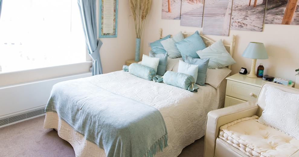 A tranquil double bedroom, the white metal framed double bed sits centrally on the back wall against the white décor. The pale blue curtains add a calming colour to the room. The white bedside tables either side of the bed are adorned with a teal lamp and ceramic vase.