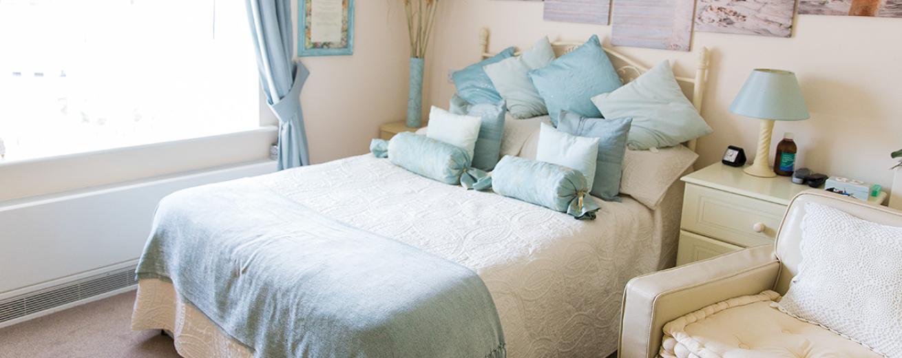 A tranquil double bedroom, the white metal framed double bed sits centrally on the back wall against the white décor. The pale blue curtains add a calming colour to the room. The white bedside tables either side of the bed are adorned with a teal lamp and ceramic vase.