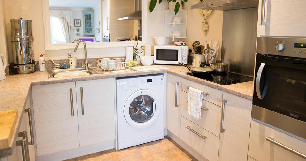 A compact white kitchen, featuring light beach wood cabinetry with white painted walls. The backsplash, wall mounted oven and extractor fan and sink are all in chrome which reflects the light perfectly. The room is warmed by a mottled kitchen counter surrounding the room, broken up by the electric hob, baking accessories used as decorations and a washing machine.
