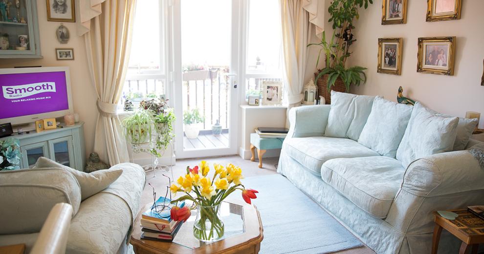 A cosy living area housing a floral patterned pale blue three seater sofa and matching arm chair both facing a television in the corner of the room sat on top of a pale blue painted television unit. A patio door leads out onto a private balcony area. Arrays of different sized plants are located around the room providing a homely charm.