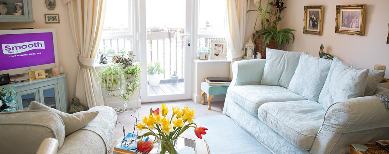 A cosy living area housing a floral patterned pale blue three seater sofa and matching arm chair both facing a television in the corner of the room sat on top of a pale blue painted television unit. A patio door leads out onto a private balcony area. Arrays of different sized plants are located around the room providing a homely charm.
