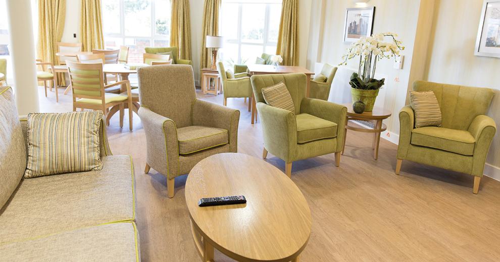 A airy communal seating area. Displaying a soft cream patterned design feature wall. The light wooden oval coffee tables and circular side table complement the green sofas and armchairs. Matching dining seating areas are tucked in just behind the comfy chairs.