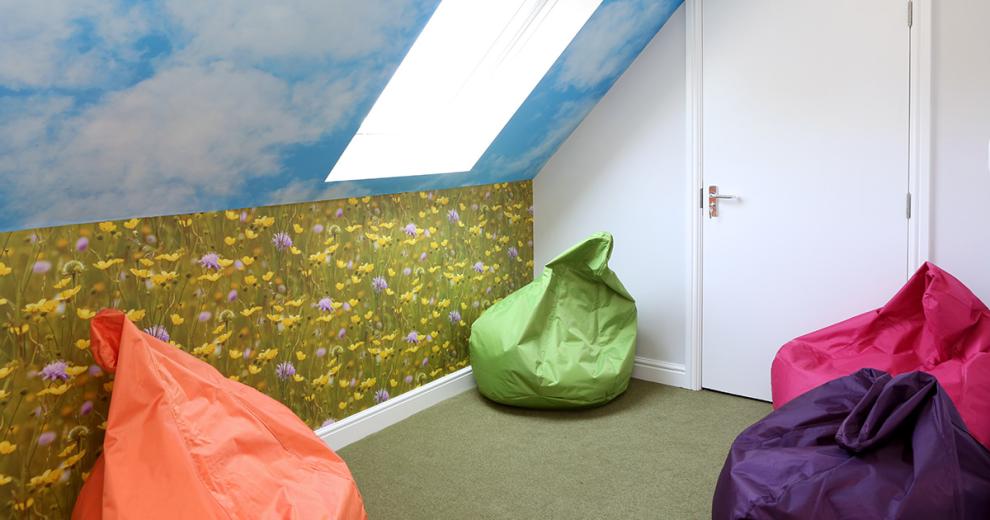Green, orange, pink and purple bean bags places in a cosy angled room decorated with calming outdoor wallpaper, showcasing a blue sky and wildflower field. As well as having a large sky light to add to the outdoor nature theme.