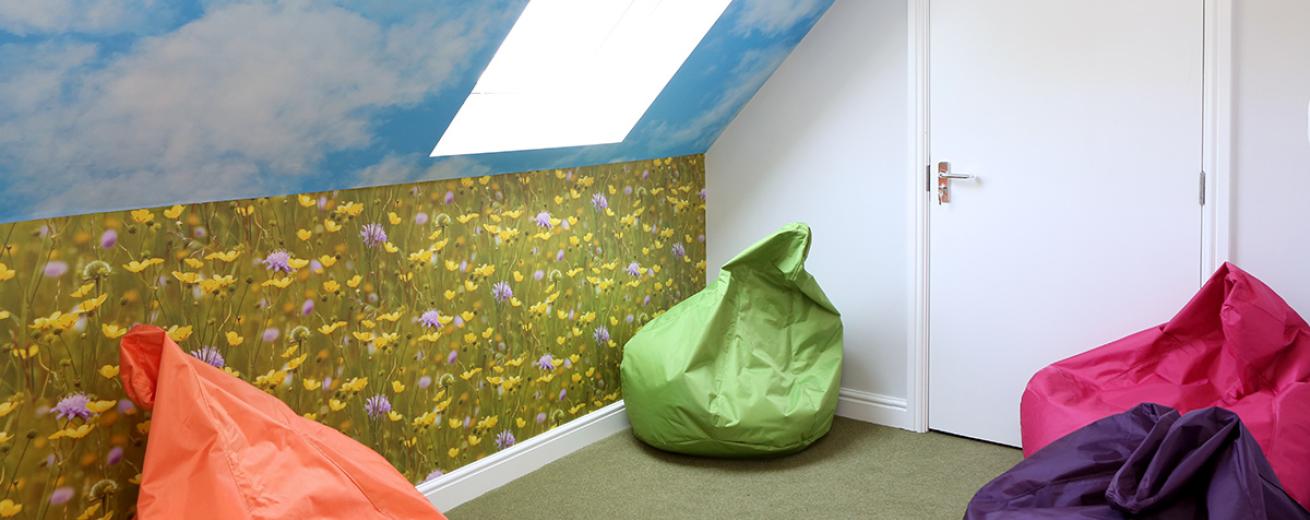 Green, orange, pink and purple bean bags places in a cosy angled room decorated with calming outdoor wallpaper, showcasing a blue sky and wildflower field. As well as having a large sky light to add to the outdoor nature theme.