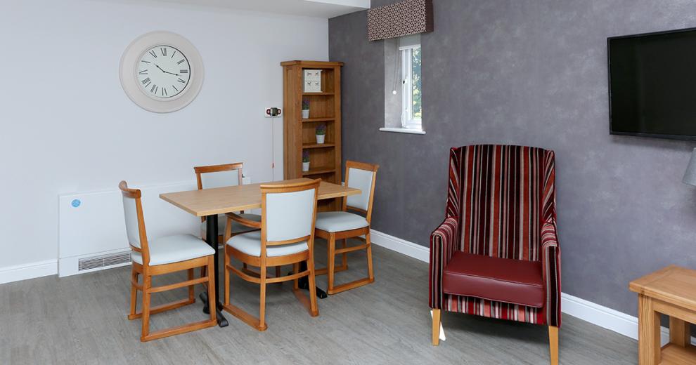 Grey mottled wallpapered seating area consisting of a four-person seated dining table and burgundy toned striped armchair around a wall mounted television.