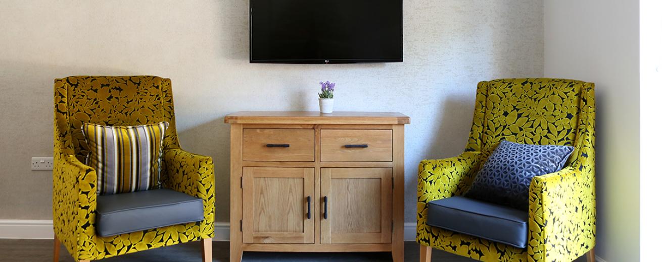Beautiful yellow and navy fabric armchairs placed around a pine television unit with black handles that compliments the wall mounted television above it.