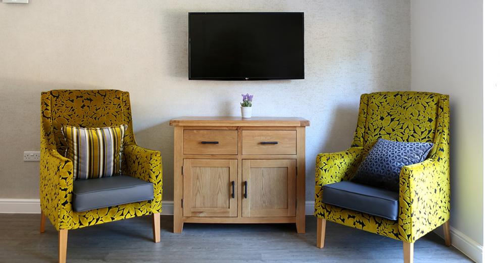 Beautiful yellow and navy fabric armchairs placed around a pine television unit with black handles that compliments the wall mounted television above it.