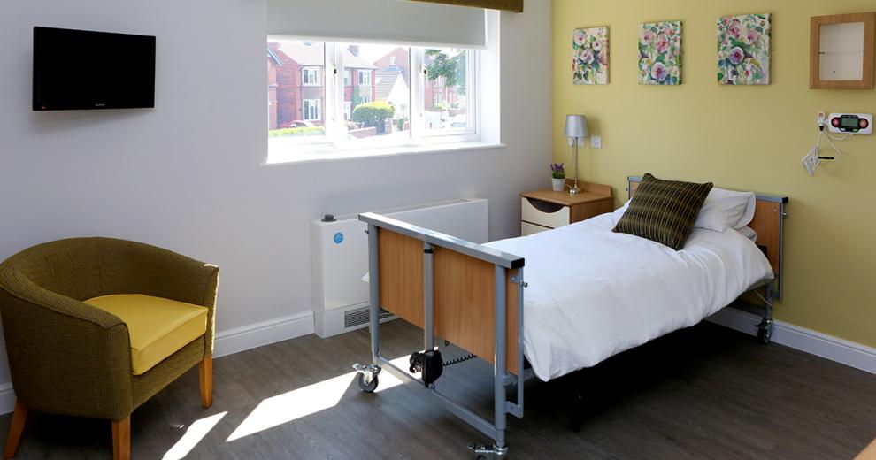 Assisted living single bed room, using a pastel yellow and floral accessories allowing some of the outside in. The room also has a small bucket armchair and wall mounted television.