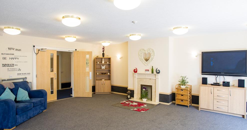 Well lit, considerable sized communal living area. With simplistic white painted walls and feature black stripe across the bottom. The decorative fireplace is the focal point of the room. A blue suede sofa is angled towards a large, mounted television which sits above a television unit that matches the stand-alone shelving unit.