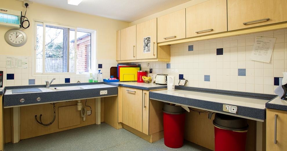 A fair sized cream kitchen with plenty of upper light wooden cabinetry as well, height adjustable work surfaces and sink area for easy accessibility. The bold colours for the chopping boards, bread bin, bins and toaster add personality to the room.