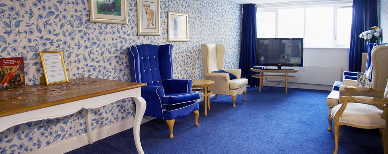 A blue regal room, with bold blue carpets, blue and white patterned wallpaper a large television. Two royal blue chesterfield arm chairs with gold trim are placed against the wall. As well as two cream arm chairs. A large ornate table homes a half complete jigsaw puzzle.