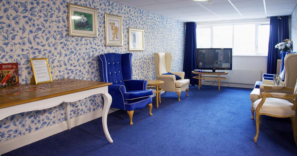 A blue regal room, with bold blue carpets, blue and white patterned wallpaper a large television. Two royal blue chesterfield arm chairs with gold trim are placed against the wall. As well as two cream arm chairs. A large ornate table homes a half complete jigsaw puzzle.