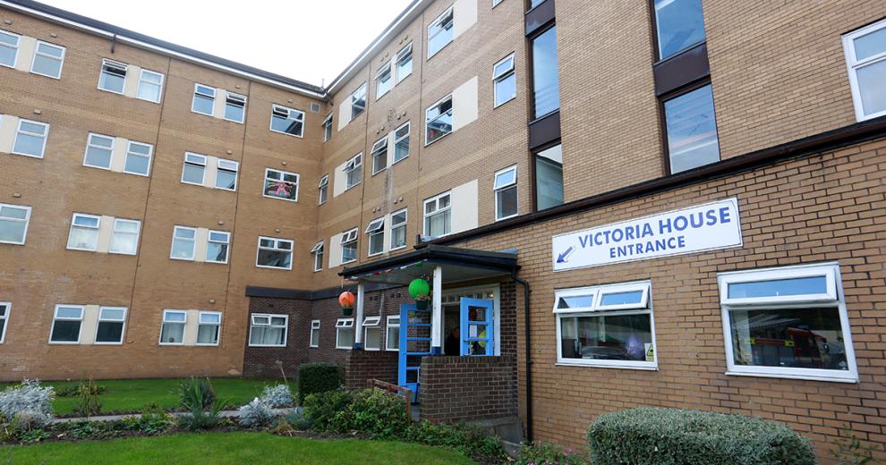 Four storey apartment building, with vivid blue doors to enter decorated with orange and green lanterns to create an inviting entrance. A large grass front garden lined with shrubbery.