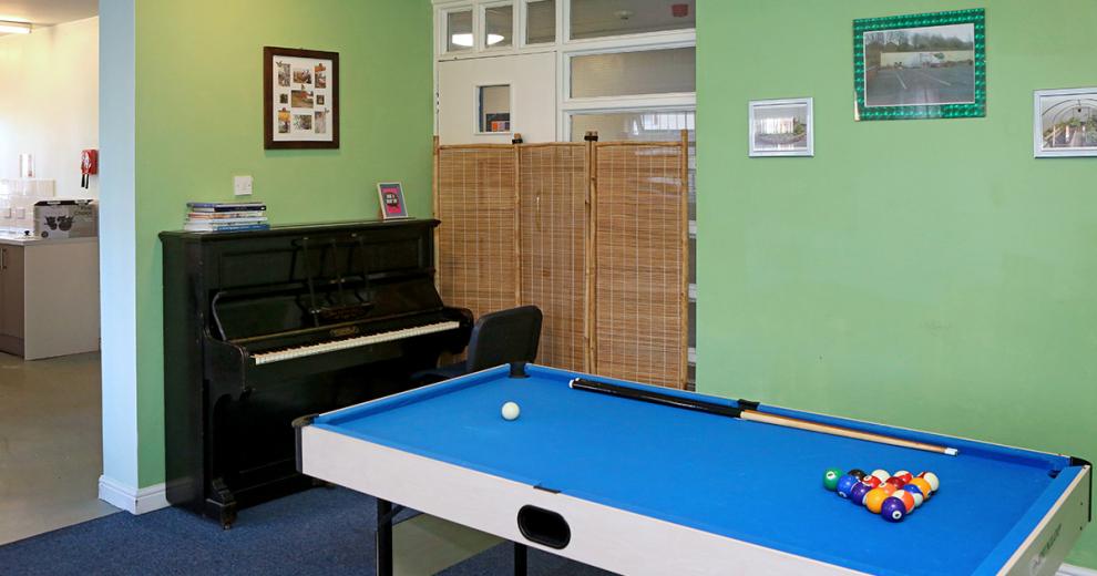 Small collapsible blue, white and black set up snooker table, in a soothing green painted room with personal photographs on the wall. The grand black piano fills the wall space offered next to the opening for the kitchen.