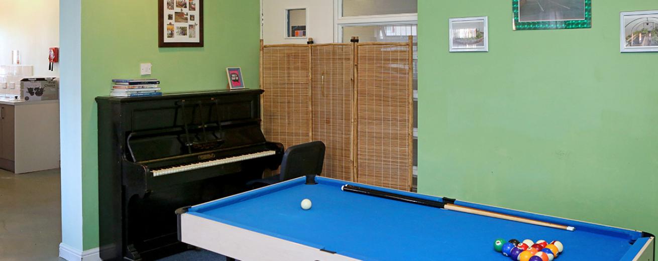 Small collapsible blue, white and black set up snooker table, in a soothing green painted room with personal photographs on the wall. The grand black piano fills the wall space offered next to the opening for the kitchen.